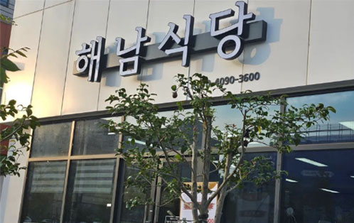 해남식당 건물 외부 전경. 흰색 외벽에 검은색 입체 글자로 ‘해남식당’ 간판이 붙어 있고, 아래에는 전화번호 4090-3600이 표시되어 있다. 건물 앞에는 작은 나무가 자라고 있으며, 유리창 너머로 식당 내부의 조명이 보인다.