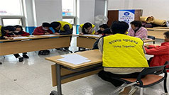 동신대학교 작업치료학과 조끼를 입고 있는 학생들과 책상 앞에 둘러 앉아서 종이에 글씨를 쓰고 있는 노인들 모습