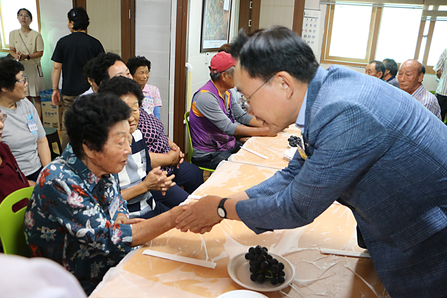 무더위 쉼터 현장 점검 나주시장이 마을 경로당을 방문하여 입식테이블에 앉아 있는 여성 어르신의 한손을 두손으로 잡으며 악수를 나누는 모습