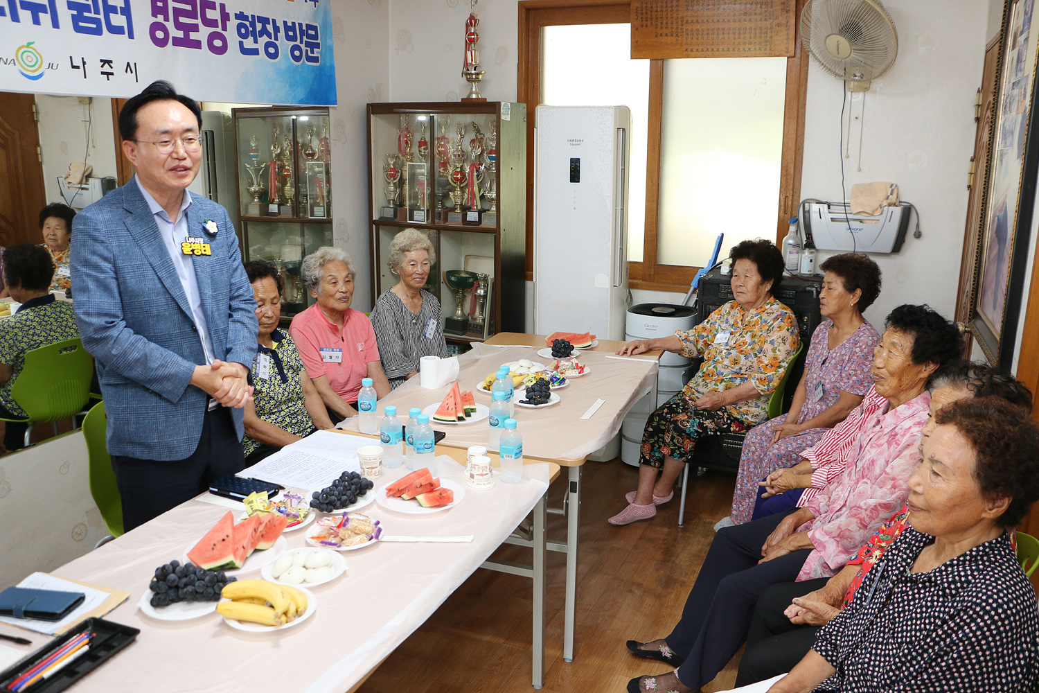 무더위 쉼터 현장 점검 나주시장이 두손을 배꼽 정도에 모으고 마을 어르신들께 인사말을 하고 있는 모습과 자리에 앉아 있는 마을 어르신들의 모습