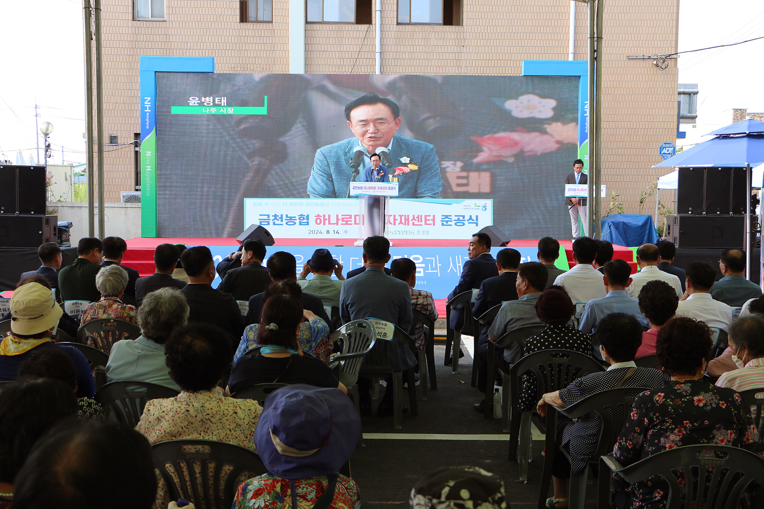 금천농협 하나로마트 자재센터 준공식 나주시장이 무대 위 단상에서 축사를 하고 있는 모습과 참석한 면민들의 앉아 있는 뒷모습