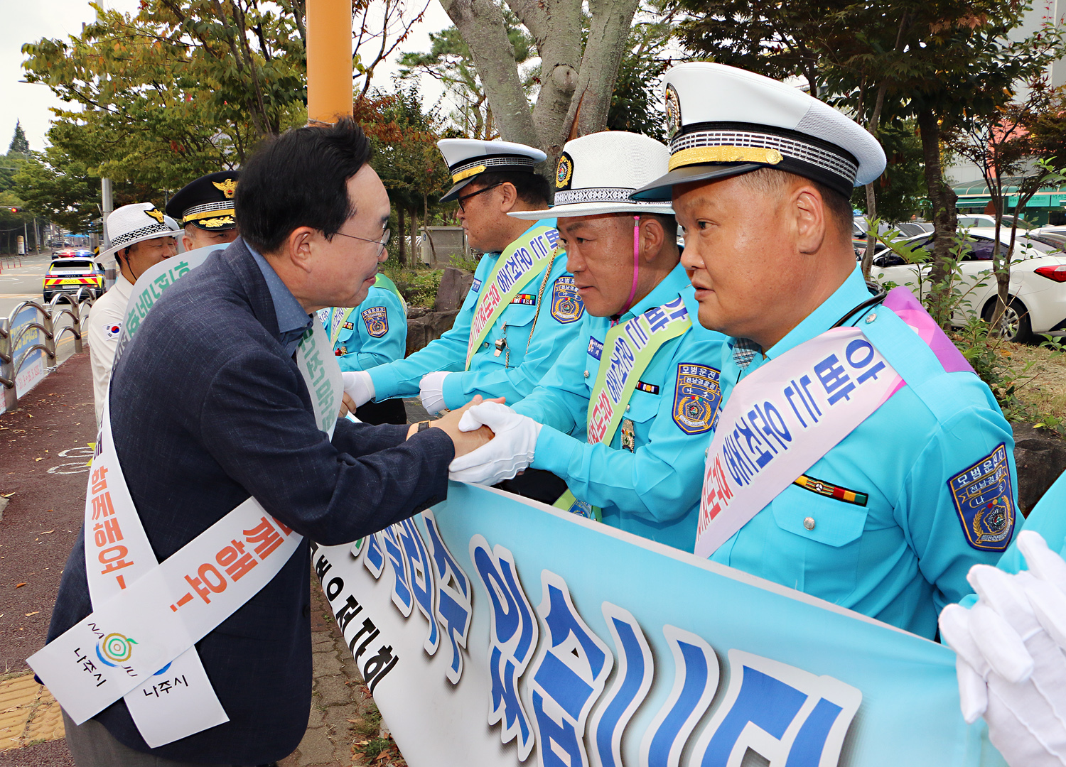 민, 관, 경 합동 어린이 교통안전 캠페인 나주시장이 현수막을 펼치고 캠페인을 벌이고 있는 모범운전자회 남성 회원분과 두손을 맞잡으며 인사를 하고 있는 모습