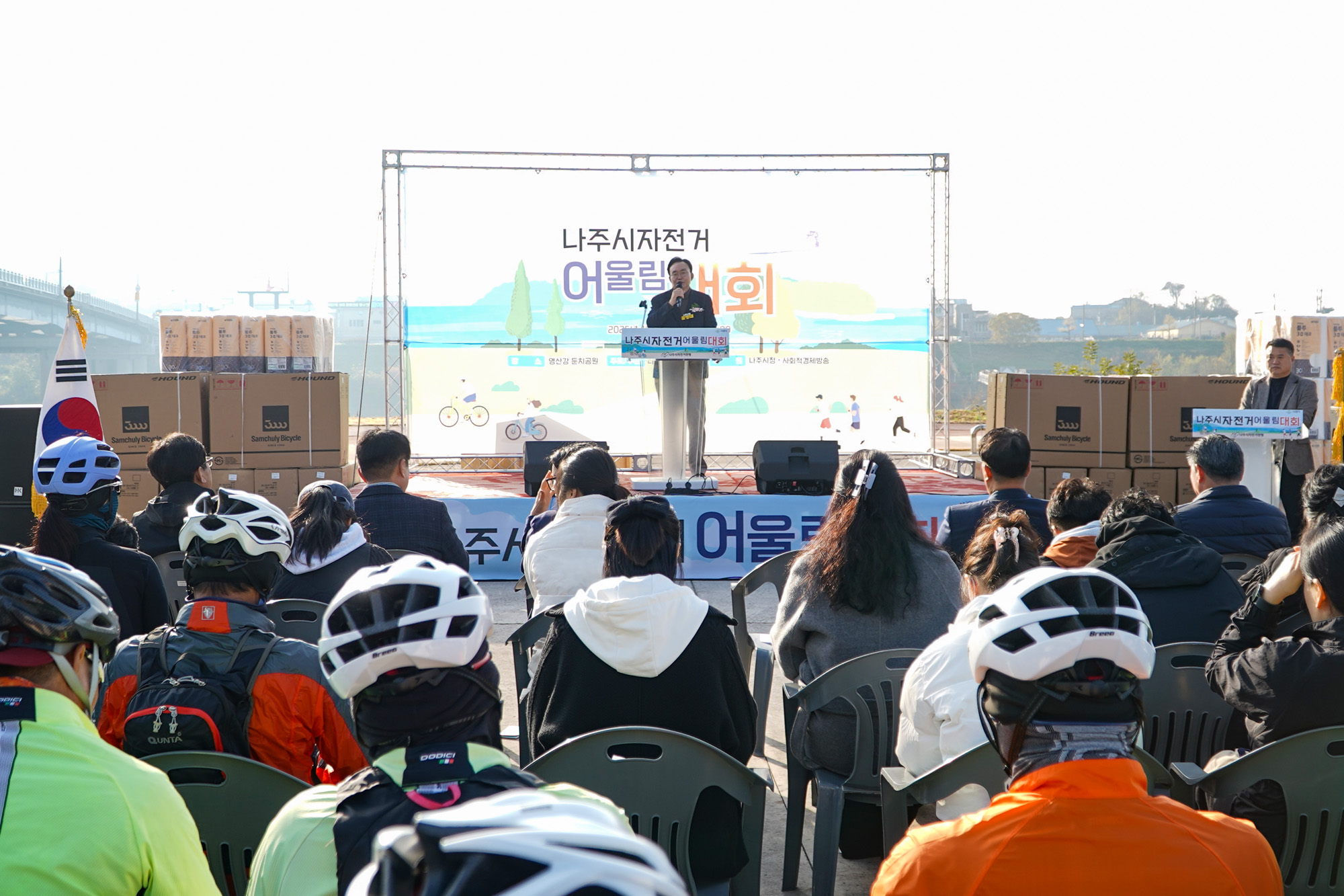  '나주시자전거 어울림대회' 개회식 현장. 무대 위의 연설자(나주시장 윤병태)를 향해 자전거 헬멧을 쓴 참가자들이 의자에 앉아 경청하고 있는 모습