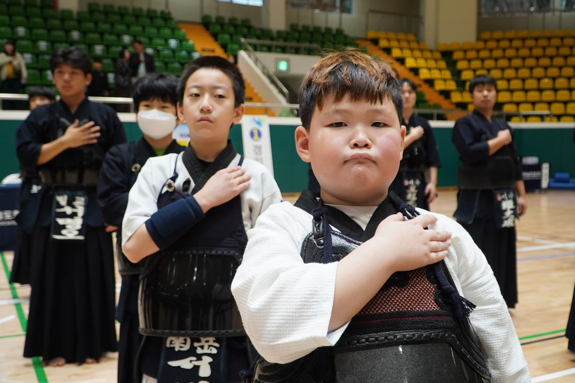 검도복과 호구를 착용한 소년들이 체육관에서 국기에 대한 경례를 하듯 가슴에 손을 얹고 서 있다.