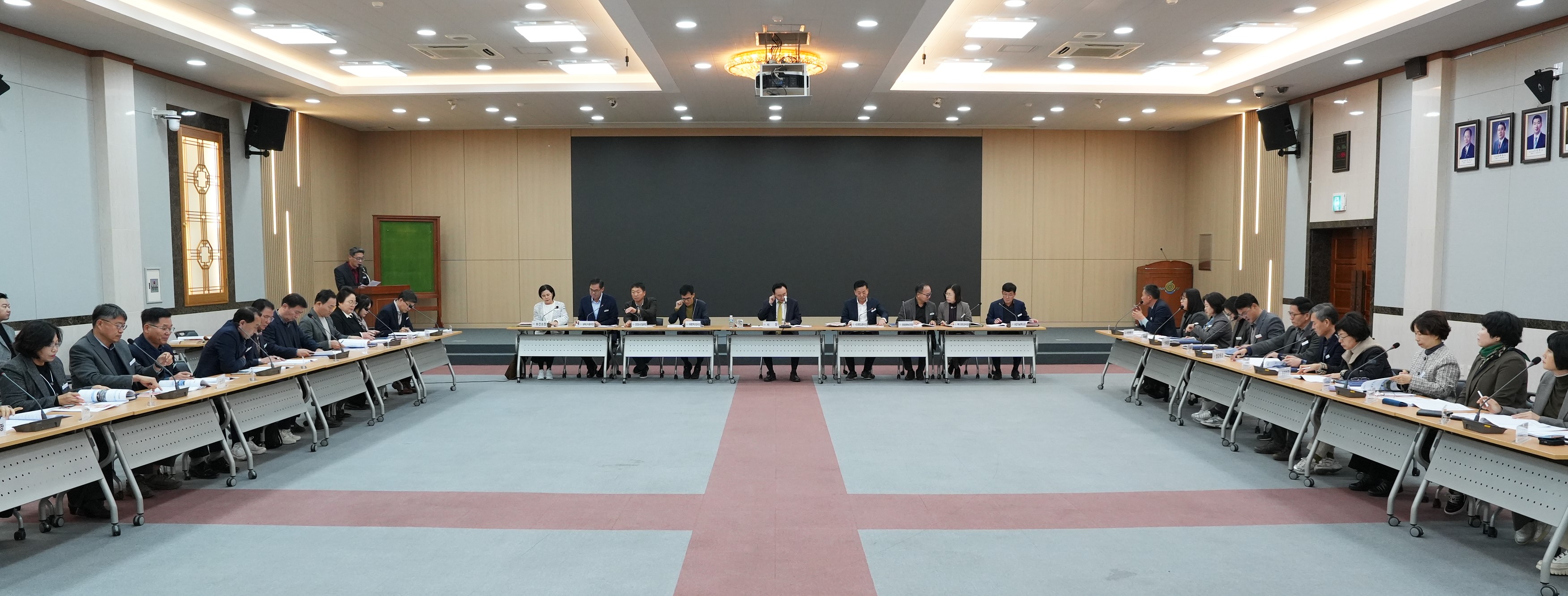  넓은 회의장에서 'ㄷ'자 형태로 테이블에 둘러앉아 회의하는 많은 사람들의 모습. 중앙에는 주요 관계자들이, 양 옆 긴 테이블에는 여러 참석자들이 앉아있다. 뒤쪽 벽에는 검은색 스크린과 액자들이 걸려있다.