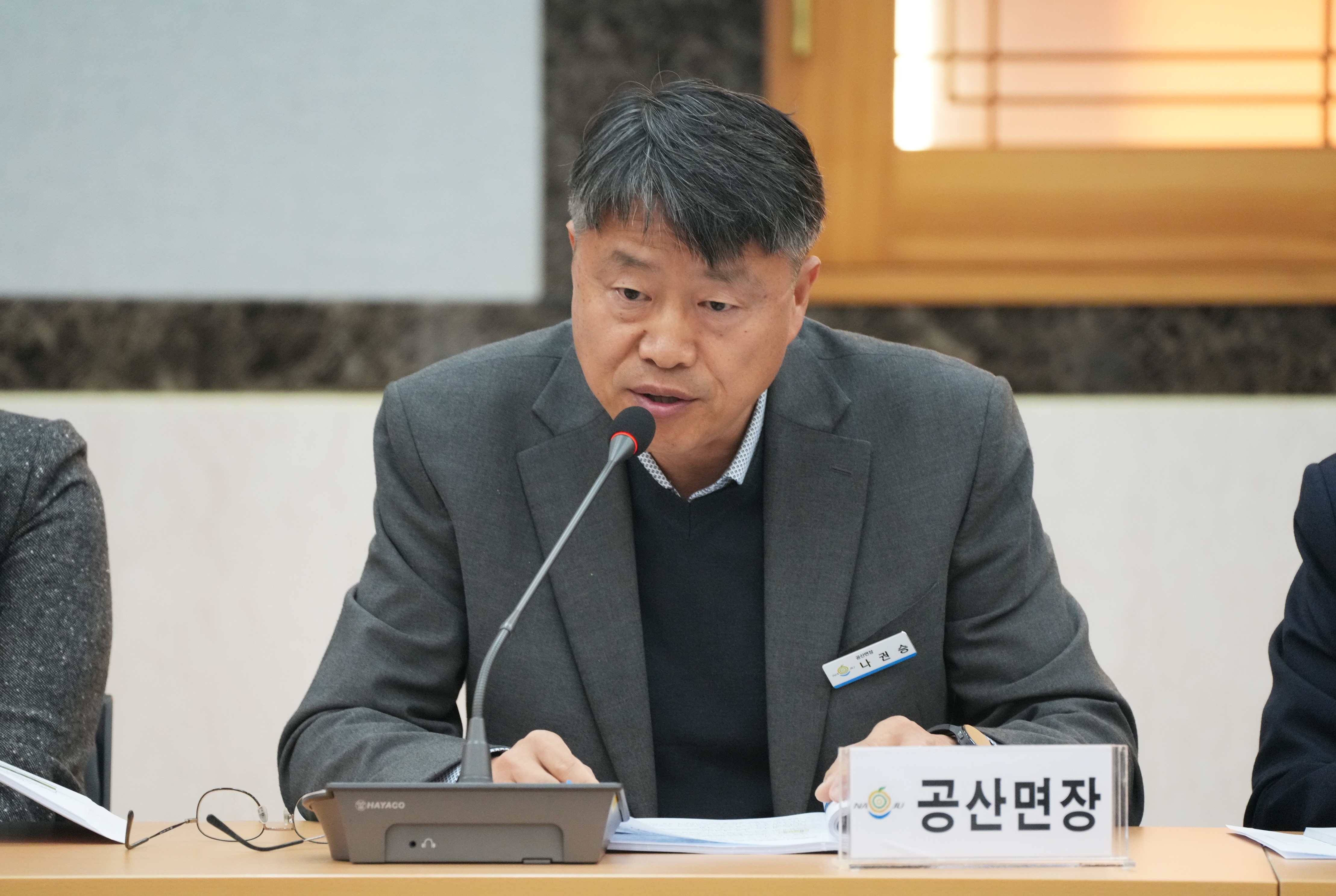  회의에 참석한 중년 남성이 '공산면장' 명패를 앞에 두고 마이크를 잡고 발언하는 모습. 짙은 회색 재킷을 입고 있다.