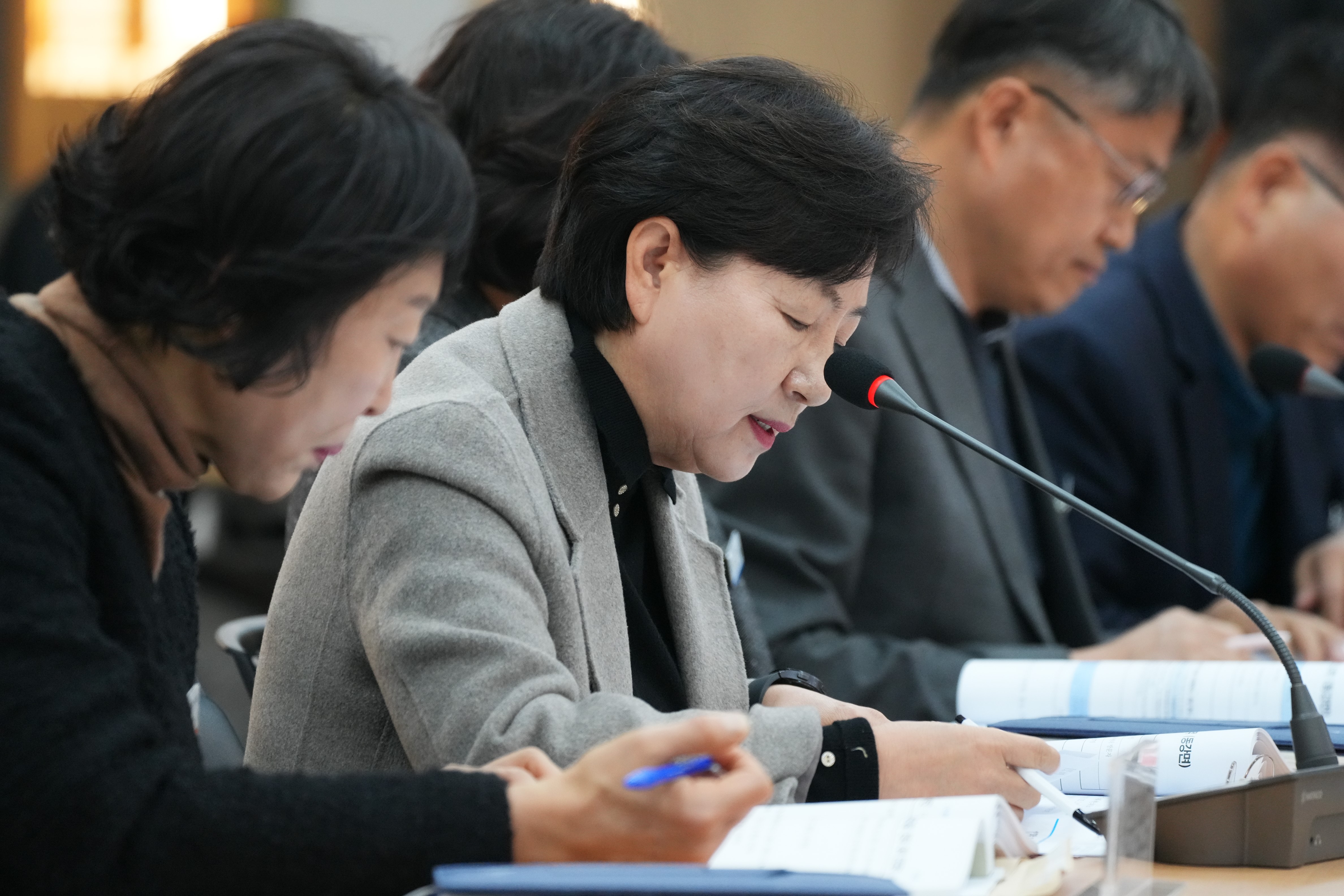  회의에 참석한 한 여성이 마이크 앞에서 고개를 숙여 서류를 읽거나 메모를 하고 있는 모습. 회색 재킷을 입고 있다.