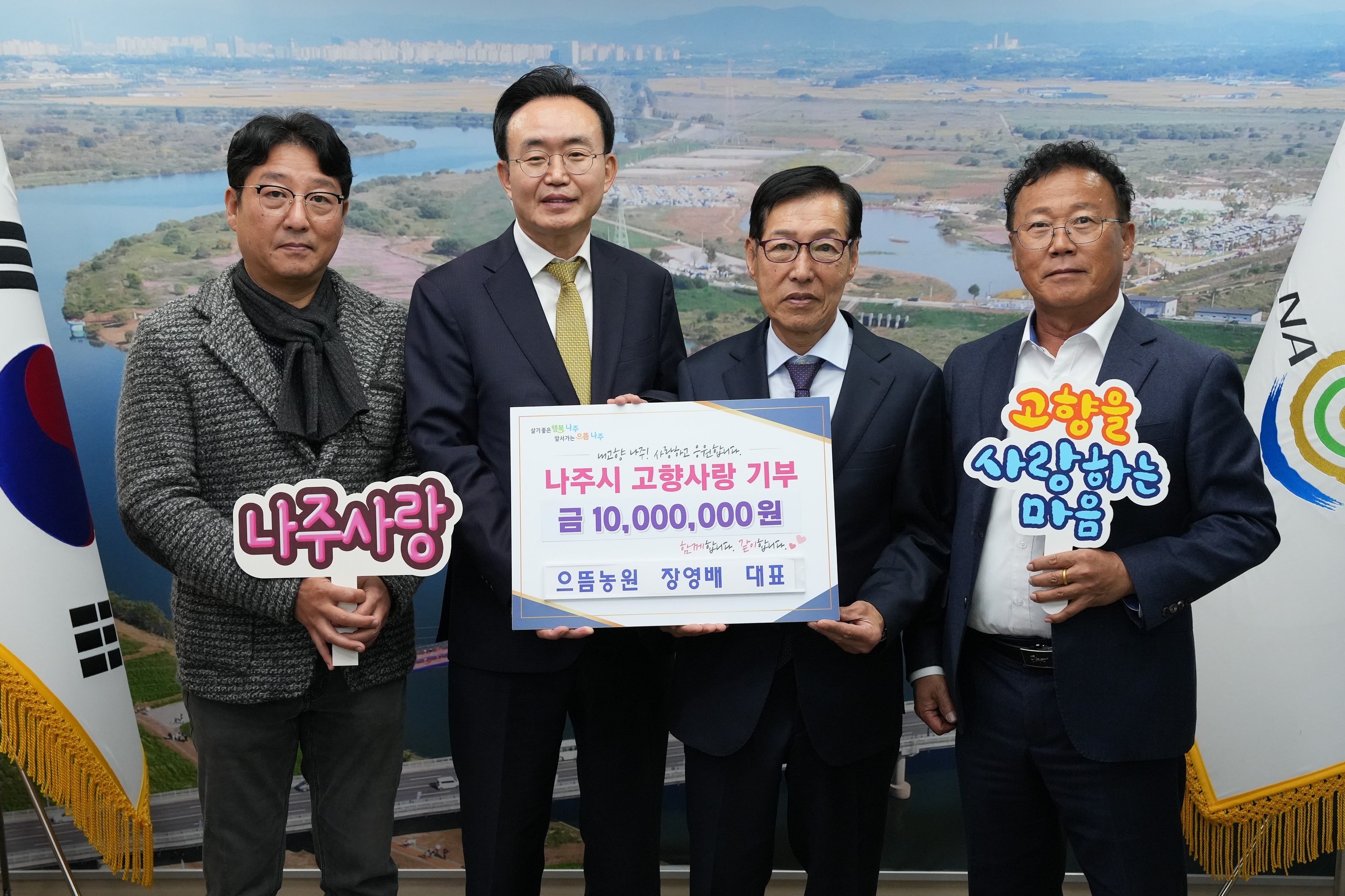 야외 풍경을 배경으로 네 명의 남성(가운데 나주시장 윤병태)이 서서 사진을 찍고 있다. 중앙의 두 남성은 '나주시 고향사랑 기부 금 10,000,000원'이 적힌 기부증서를 들고 있으며, 양쪽 끝의 남성들은 '나주사랑'과 '고향을 사랑하는 마음' 문구가 적힌 손팻말을 들고 있다. 모두 미소를 짓고 있다.