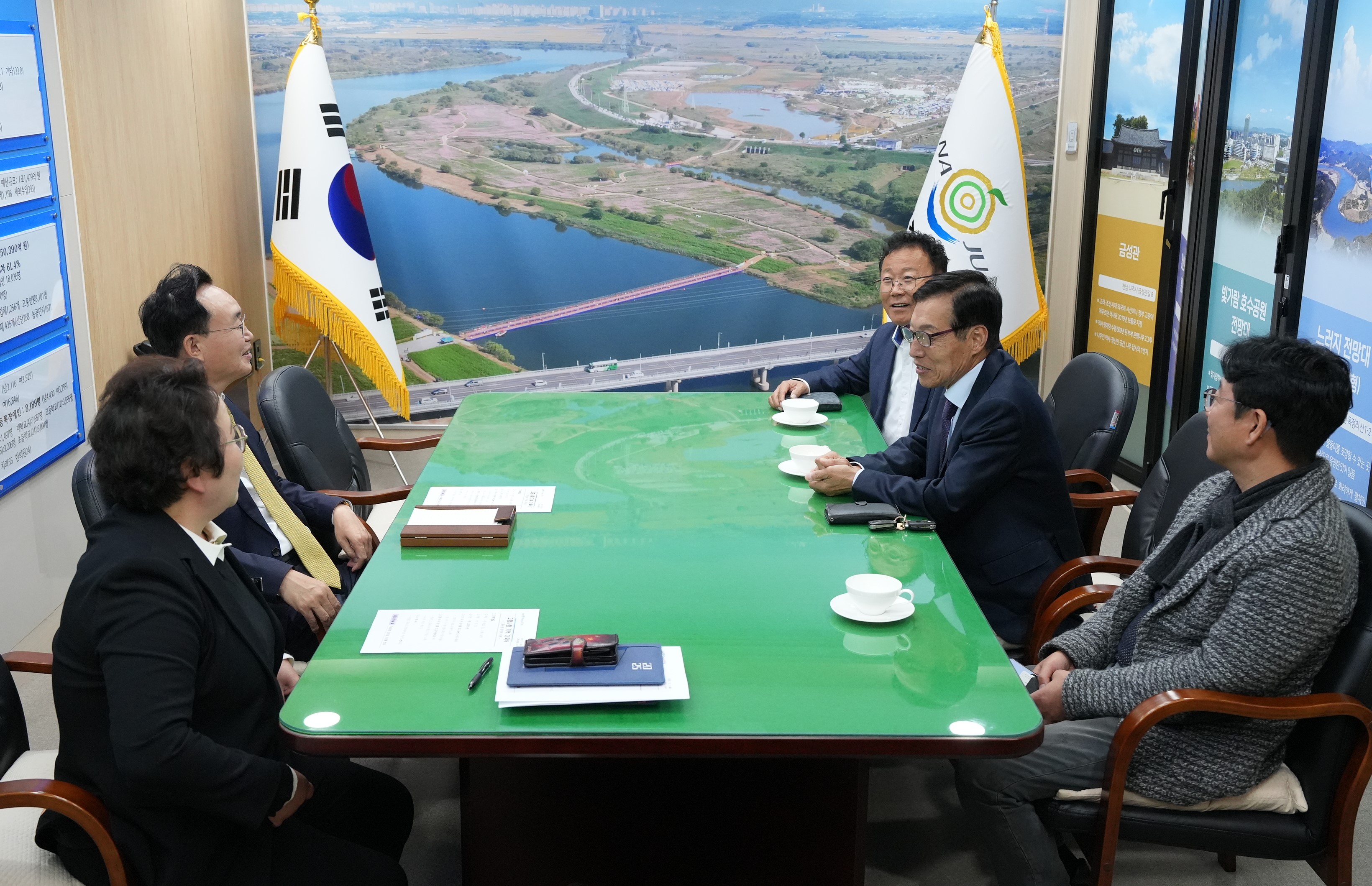 다섯 명의 남성(나주시장 윤병태 포함)이 큰 녹색 테이블에 둘러앉아 회의를 하는 모습. 정장을 입은 사람들이 보이며, 테이블 위에는 서류와 찻잔이 놓여 있다. 뒤쪽 벽에는 태극기와 나주 로고가 있는 깃발, 그리고 강이 보이는 야외 전경 사진이 걸려 있다.
