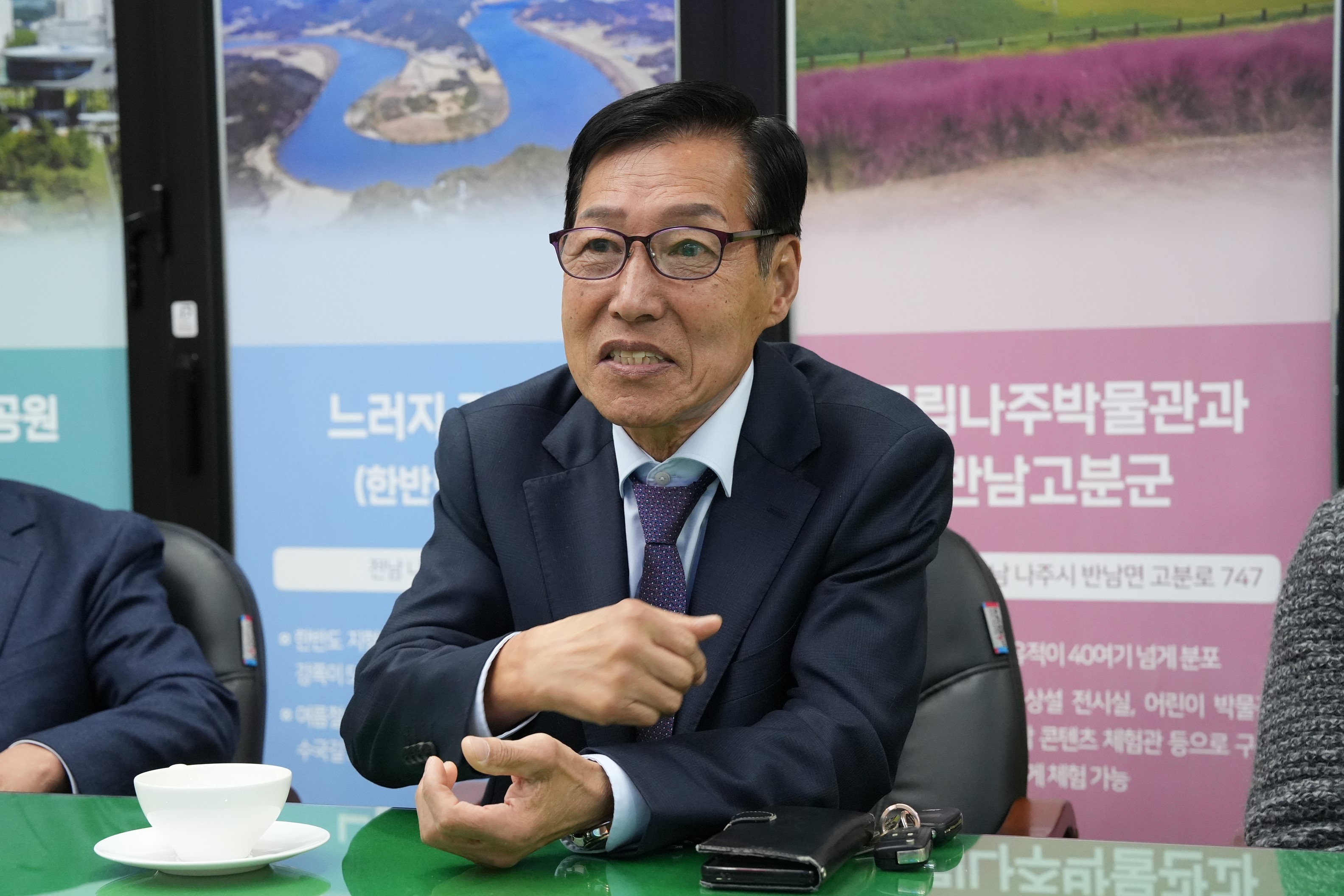 정장을 입고 테이블에 앉아 있는 중년 남성. 흰 셔츠와 짙은 색 재킷을 착용하고 있으며, 안경을 쓰고 정면을 바라보고 있다.