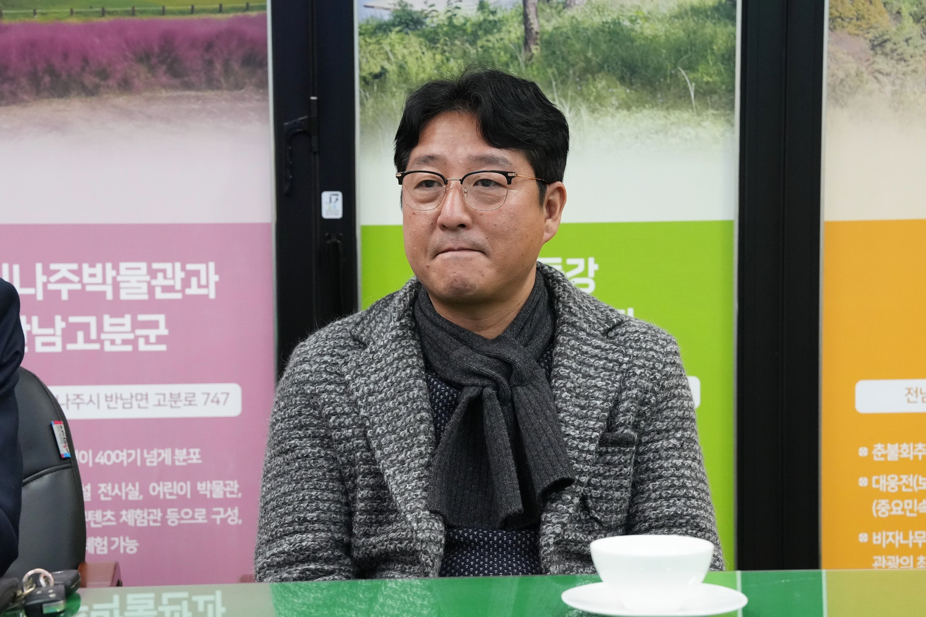 테이블에 앉아 있는 중년 남성. 목도리와 체크재킷을 착용하고 있으며, 안경을 쓰고 정면을 바라보고 있다.