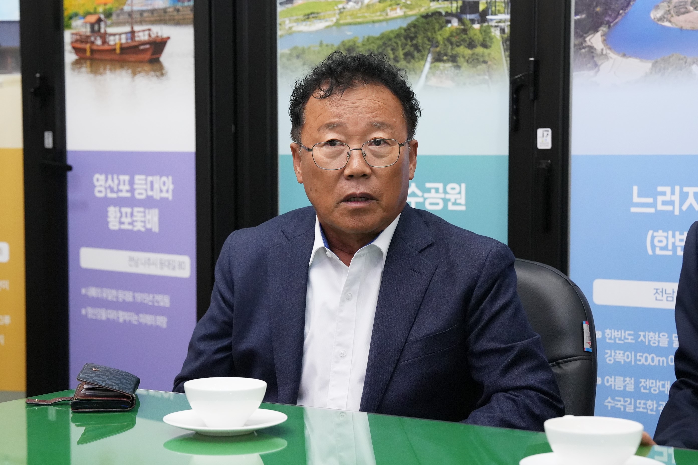 정장을 입고 테이블에 앉아 있는 중년 남성. 흰 셔츠와 남색 재킷을 착용하고 있으며, 안경을 쓰고 정면을 바라보고 있다.