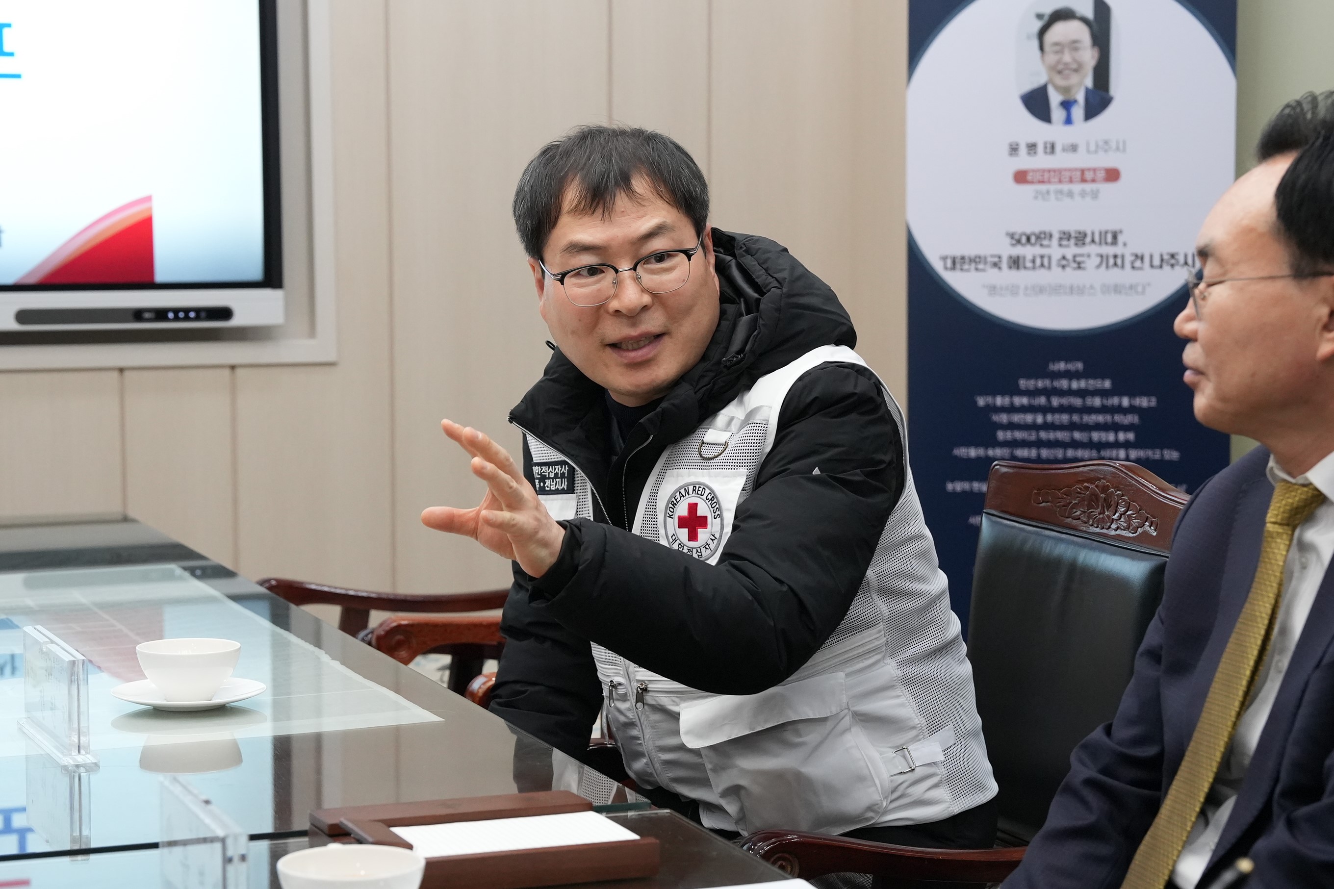 흰색 구호 조끼를 입은 중년 남성이 테이블 맞은편 사람에게 손을 사용해 설명하며 이야기하고 있는 모습