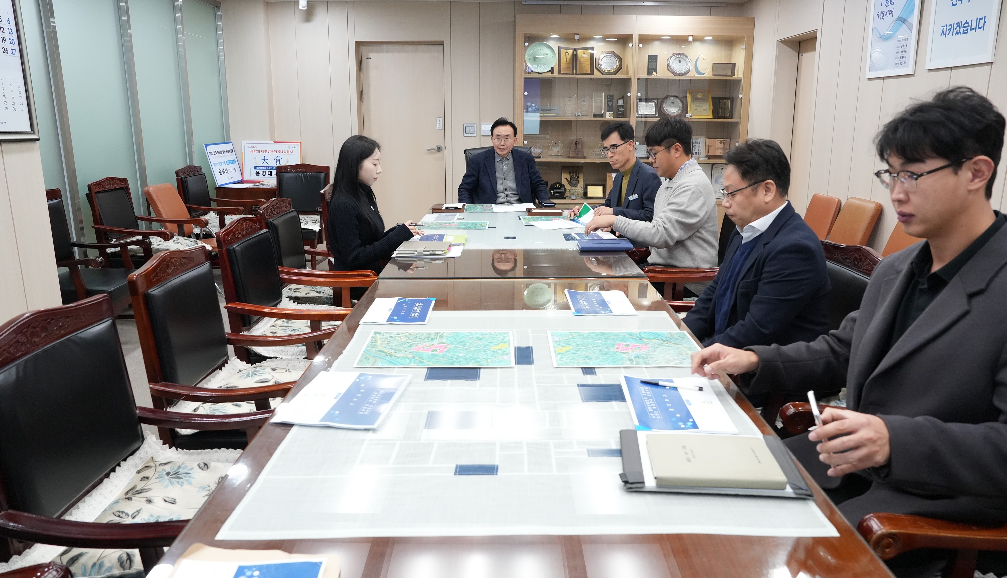  회의실 테이블에 앉아 있는 여러 명의 참석자들. 테이블 위에는 지도나 보고서가 펼쳐져 있다. 