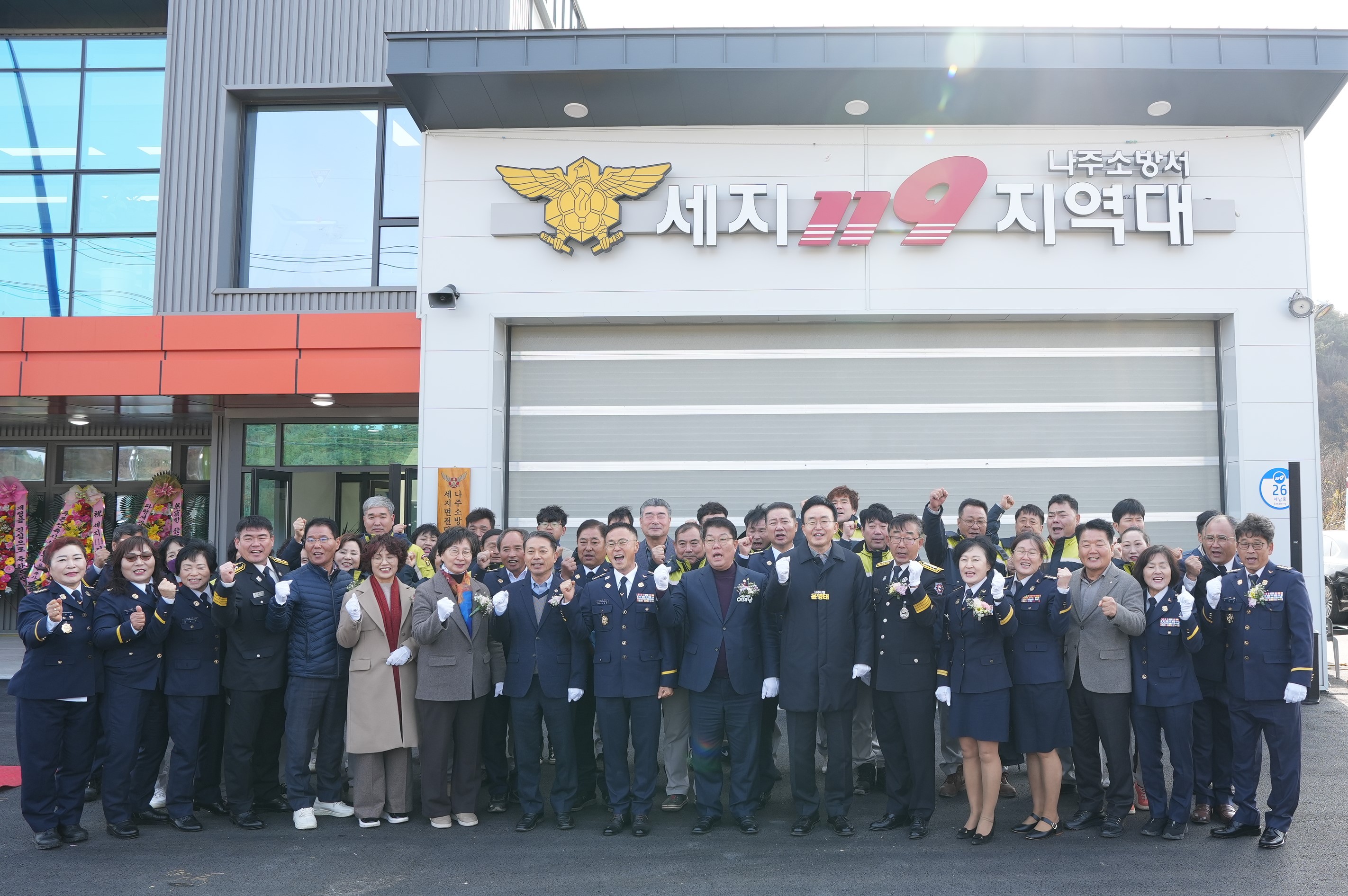  나주소방서 세지 119 지역대 개청식 단체 사진. 신축된 소방서 건물 앞에서 윤병태 나주시장(중앙)을 비롯하여 소방 관계자, 지역 인사 등 약 20여 명이 환하게 웃으며 주먹을 쥐고 기념 촬영을 하고 있다. 건물 입구 위에는 '나주소방서 세지 119 지역대' 간판이 보인다.