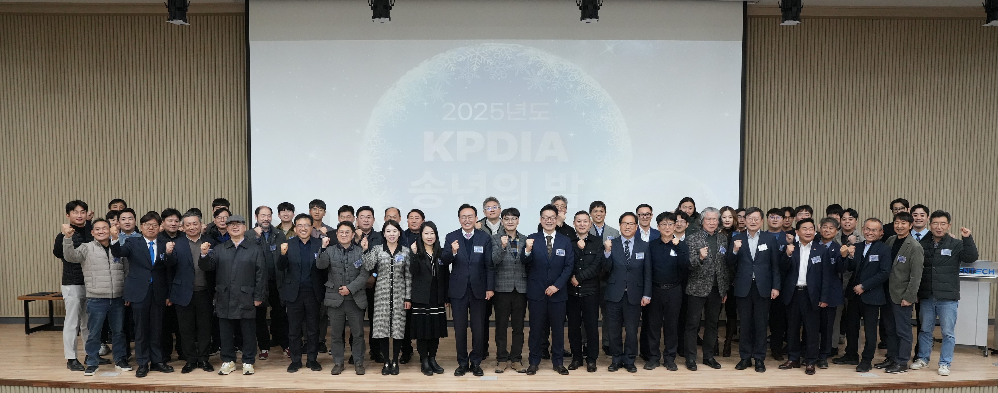  많은 인원이 행사장에서 단체 사진을 촬영하며 주먹을 쥐고 있는 모습. 배경 스크린에 '2025년도 KPDIA 송년의 밤'이라고 적혀 있음