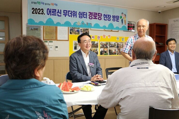우미린 경로당 무더위 쉼터 방문  경로당 어르신이 자리에 일어서서 인사말을 하고 있고 나주시장이 옆에 앉아 있는 모습