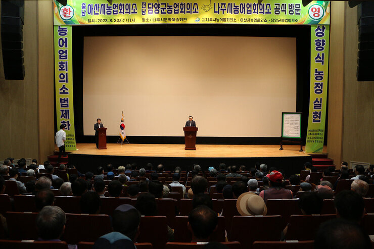 아산시, 담양군 농업회의소 나주시 방문 나주시장이 축사를 하고 있고 참석자들의 앉아 있는 모습이 보이는 전면