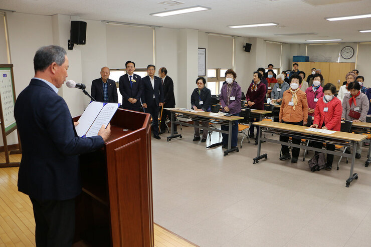 나주시 노인대학 입학식 노인대학 대표 남성 어르신의 노인강령 낭독 모습을 측면에서 촬영한 모습과 참석한 전체 어르신들이 자리에 서서 듣고 있는 모습