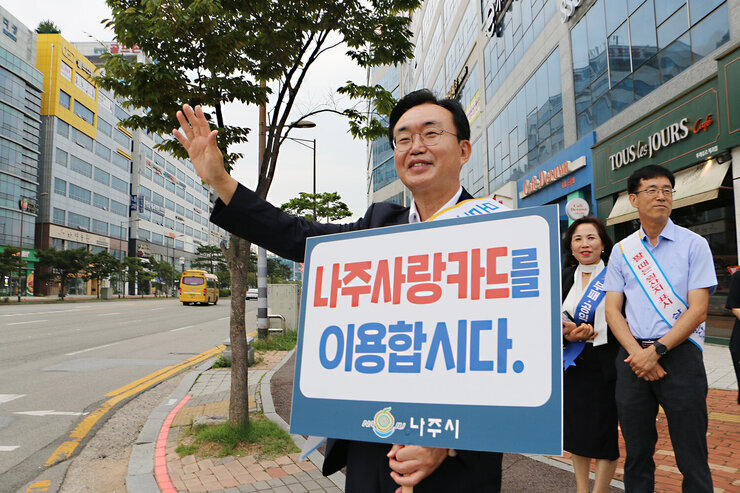 추석맞이 지역상품 판매 촉진 캠페인 및 청렴실천, 고향사랑 기부제 홍보 캠페인 나주시장이 얼굴에 미소를 지으며 나주사랑카드 이용 피켓을 들고 한손은 이동차량을 향해 손을 흔들며 홍보 캠페인을 하고 있는모습