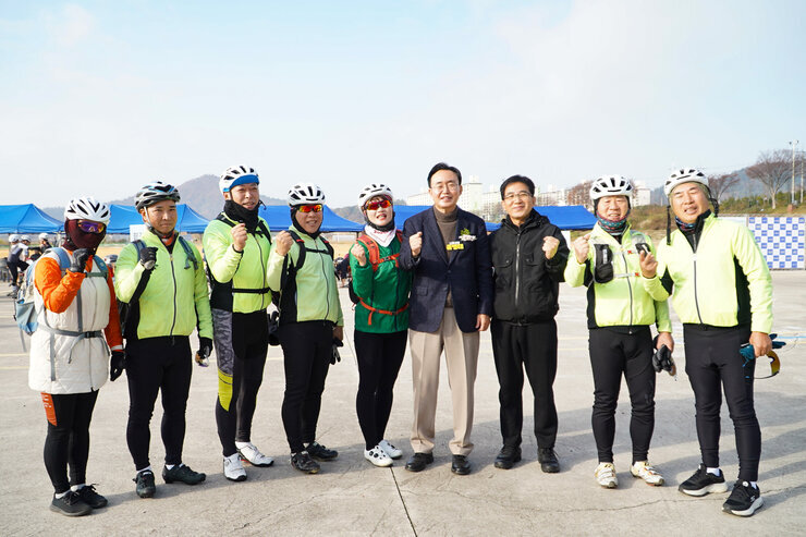 윤병태 나주시장과 밝은 색상의 자전거 복장을 갖춘 사이클 동호회 회원들 여러 명이 야외에서 함께 서서 주먹을 쥐고 단체 사진을 찍고 있다.