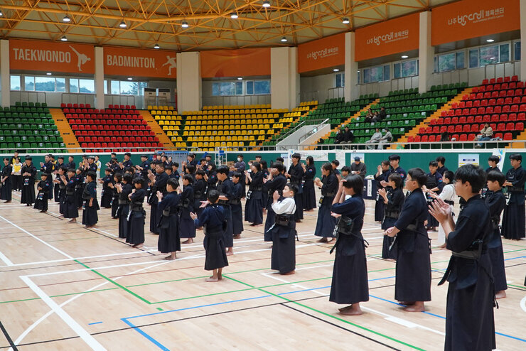 실내 체육관에서 검도복을 입은 많은 선수들이 도장 위에 서서 박수를 치고 있는 모습. 다채로운 색상의 관중석이 뒤쪽에 보인다.