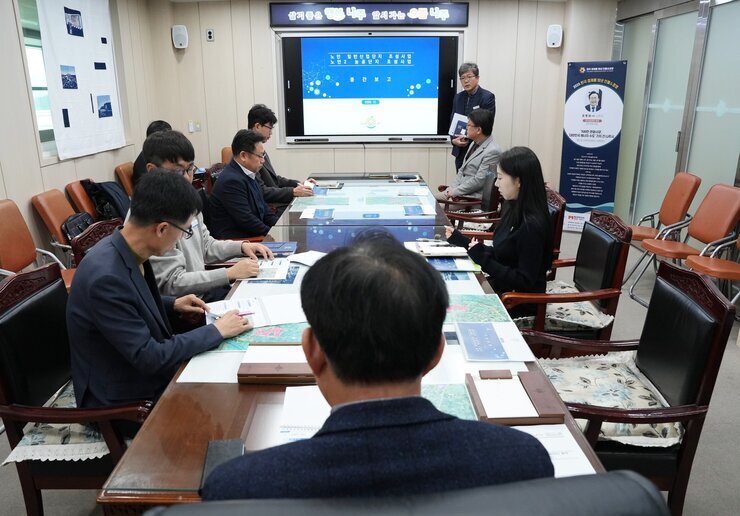  넓은 회의실에서 여러 명이 테이블을 둘러싸고 앉아 회의를 하고 있는 전경. 정면의 스크린에는 '중간보고' 관련 제목이 보이고, 한 남성이 스크린 옆에서 자료를 들고 서 있다.