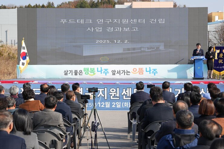  푸드테크 연구지원센터 건립공사 사업 경과보고 행사 전경. 야외 무대 위 스크린에 '푸드테크 연구지원센터 건립 사업 경과보고'와 센터 조감도가 표시되어 있다. 태극기 옆 단상에서 남녀 두 사람이 서 있으며, 앞줄에는 많은 참석자들이 의자에 앉아 경청하고 있다.