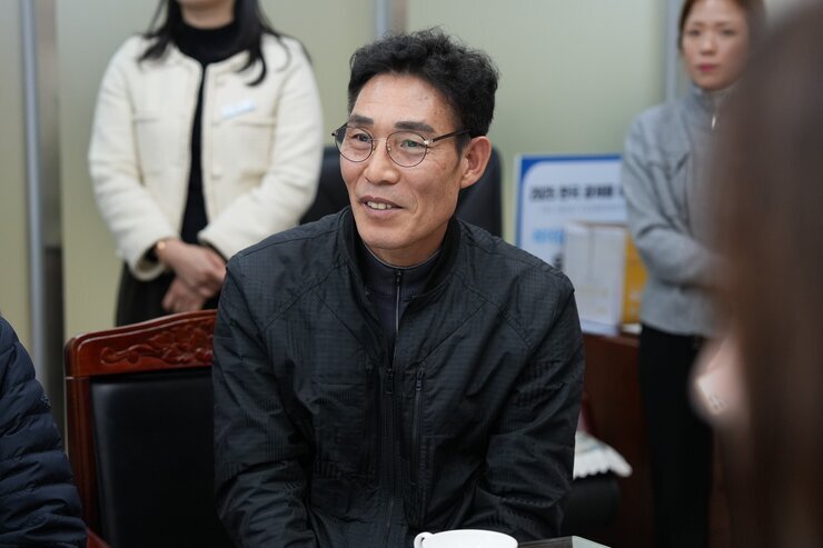  회의석상에 앉아 웃고 있는 중년 남성. 검은색 집업 재킷을 입고 안경을 쓰고 있다.