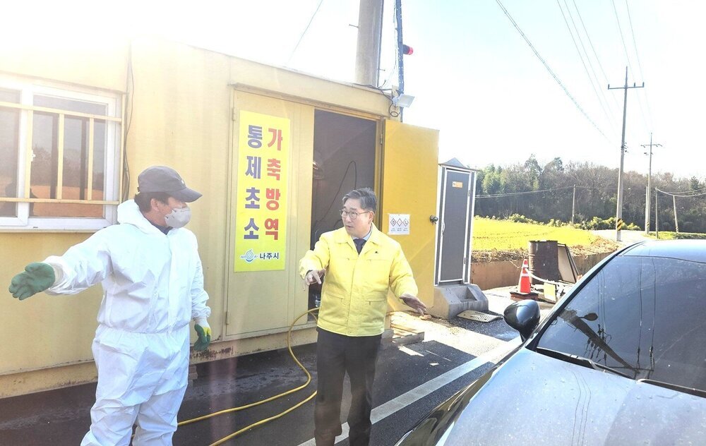 햇볕이 강한 야외에서 노란색 점퍼를 입은 한 남성이 방호복을 입은 다른 남성과 대화하고 있다. 뒤편에는 '가축방역 통제초소' 문구가 붙어있는 조립식 건물이 보이며, 앞에 검은색 차량의 일부가 보인다.