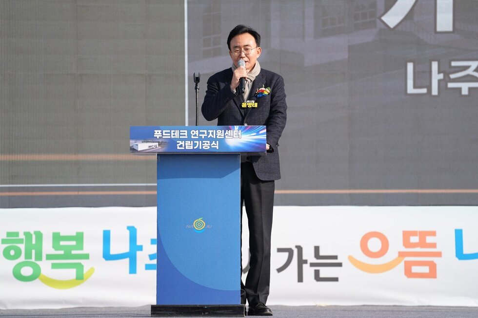 야외 행사장에서 남성 한 명(나주시장 윤병태)이 '푸드테크 연구지원센터 건립기공식' 이라고 적힌 파란색 연단에 서서 마이크를 잡고 연설하고 있는 모습. 뒤쪽에는 '나주', '행복' 등의 글자가 배경에 보인다.
