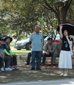 그늘에 함께 앉아있는 주민들과 서서 양산을 들고있는 여성과 몇몇 서있는 남성