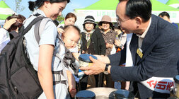 영산강축제 농업페스타 쌀소비 촉진 아침밥먹기 캠페인 나주시장이 간난아이를 몸 앞으로 맨 아이 엄마와 아이에게 노릉지 컵용기를 나눠조고 있는 모습