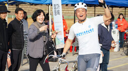  자전거 복장과 헬멧을 착용한 남성 사이클리스트가 자신의 자전거 옆에서 한 손을 들고 환하게 웃고 있으며, 옆의 여성 관람객도 웃으며 박수를 치고 있다.