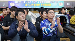  볼링 레인 앞에서 많은 사람들이 앉아 볼링 경기를 관람하고 있으며, 앞줄의 두 남성이 박수를 치고 있다. 배경에는 '제18회 나주시장기 직장인 및 클럽대항볼링대회 오신 나주시민여러분 환영합니다'라고 적힌 현수막이 걸려있다.