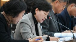  회의에 참석한 한 여성이 마이크 앞에서 고개를 숙여 서류를 읽거나 메모를 하고 있는 모습. 회색 재킷을 입고 있다.