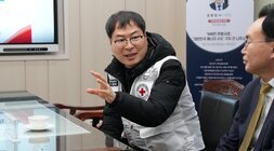  흰색 구호 조끼를 입은 중년 남성이 테이블 맞은편 사람에게 손을 사용해 설명하며 이야기하고 있는 모습