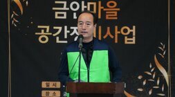  녹색 조끼를 입은 중년 남성이 단상에서 발언하는 모습. 배경에는 '금안마을 공간재생사업' 현수막이 보인다.